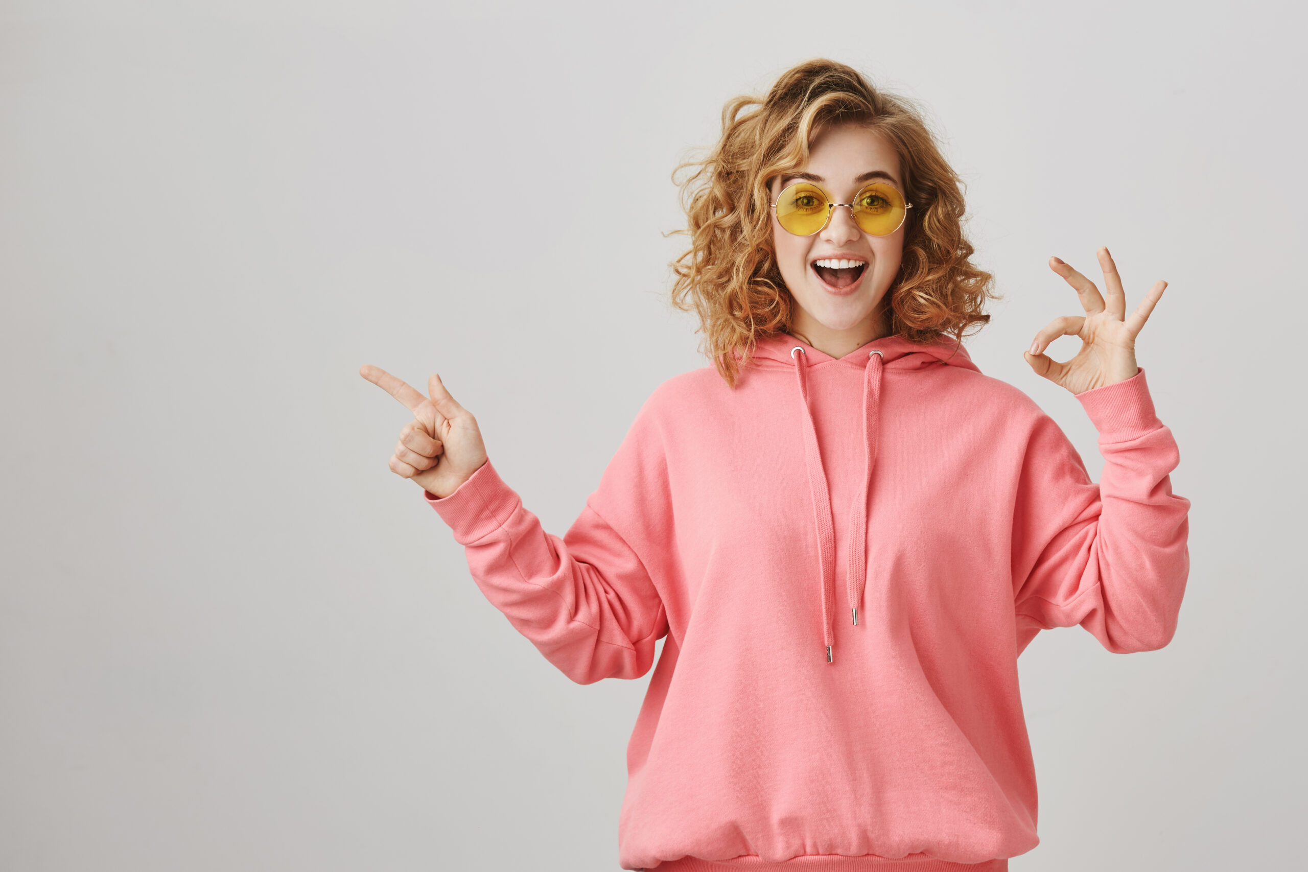 Studio portrait of emotive happy feminine woman with curly hair, wearing casual hoodie and sunglasses showing ok sign and pointing left with index finger, smiling with excitement over gray background successful habits for students